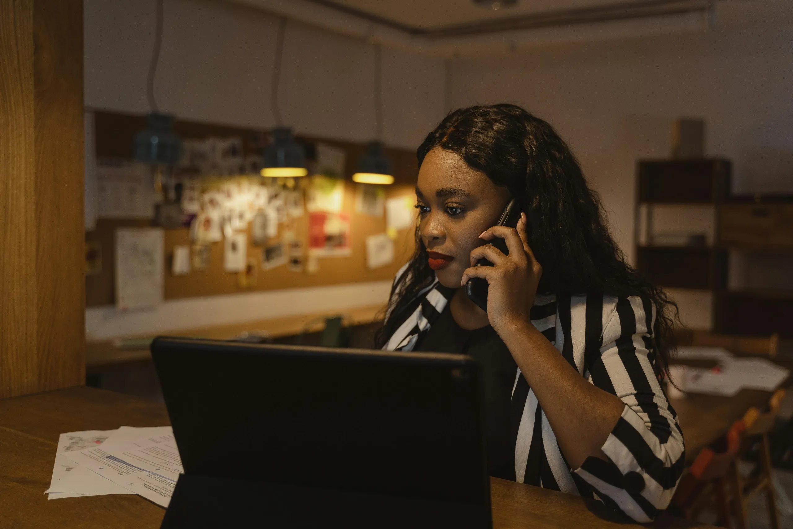 Woman in a striped blazer speaking on the phone while working on a laptop, managing documents and tasks as an online transaction coordinator.
