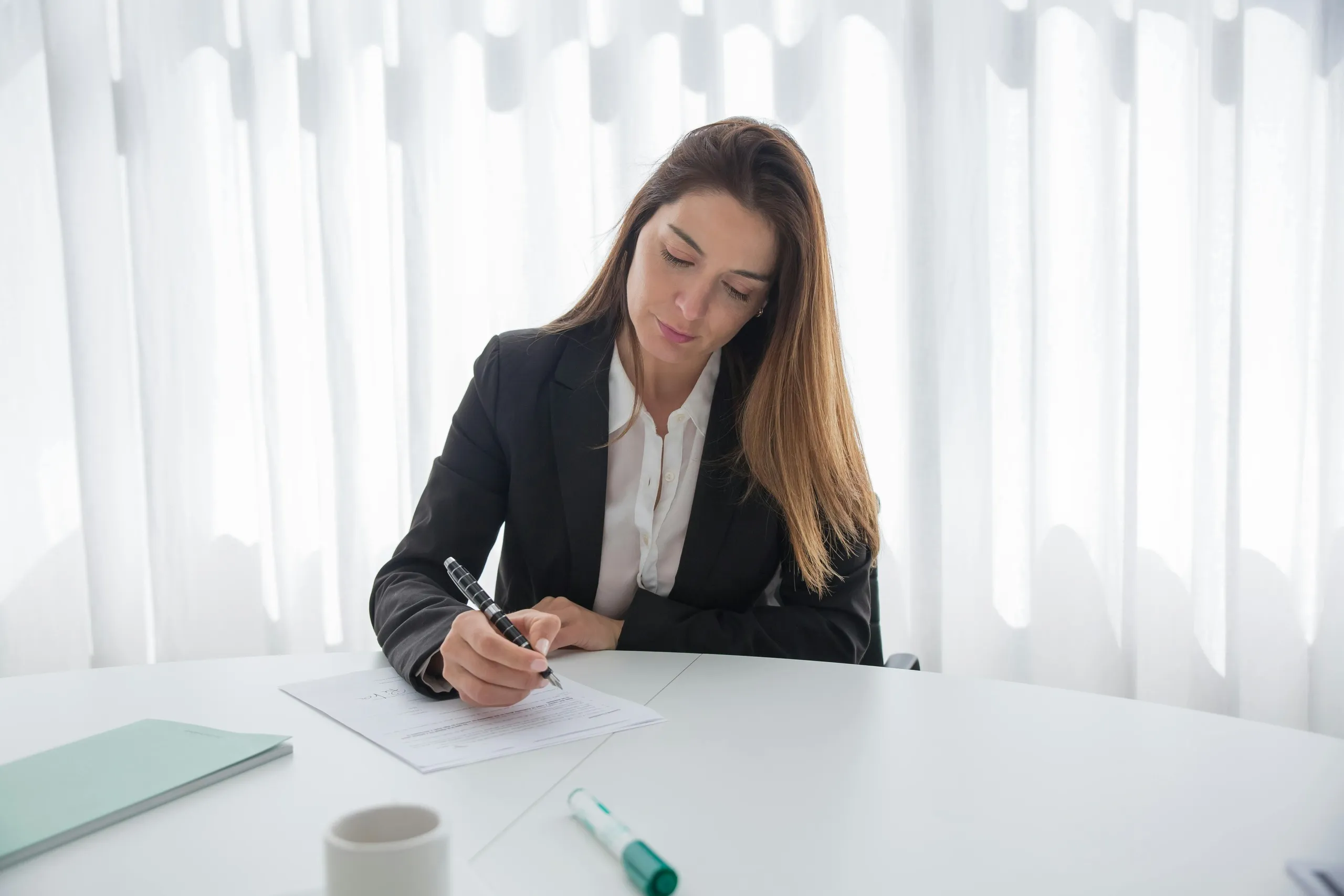 Woman in formal attire writing on documents at a desk, symbolizing remote bookkeeper skill evaluation.