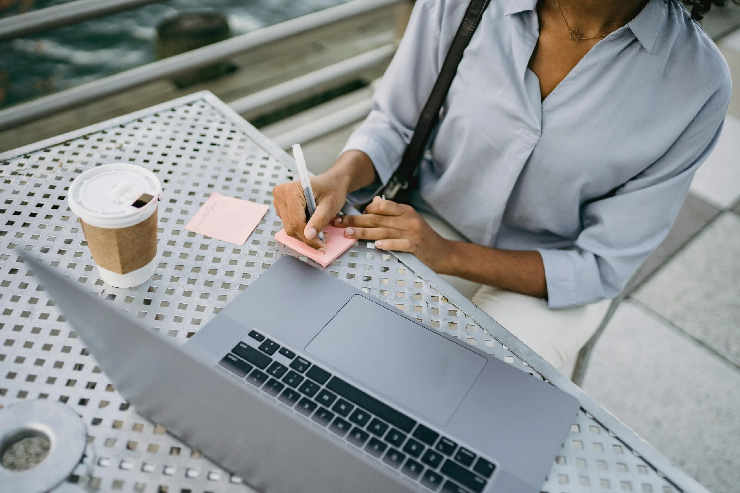 Remote executive assistant taking notes while working outdoors with a laptop and coffee.