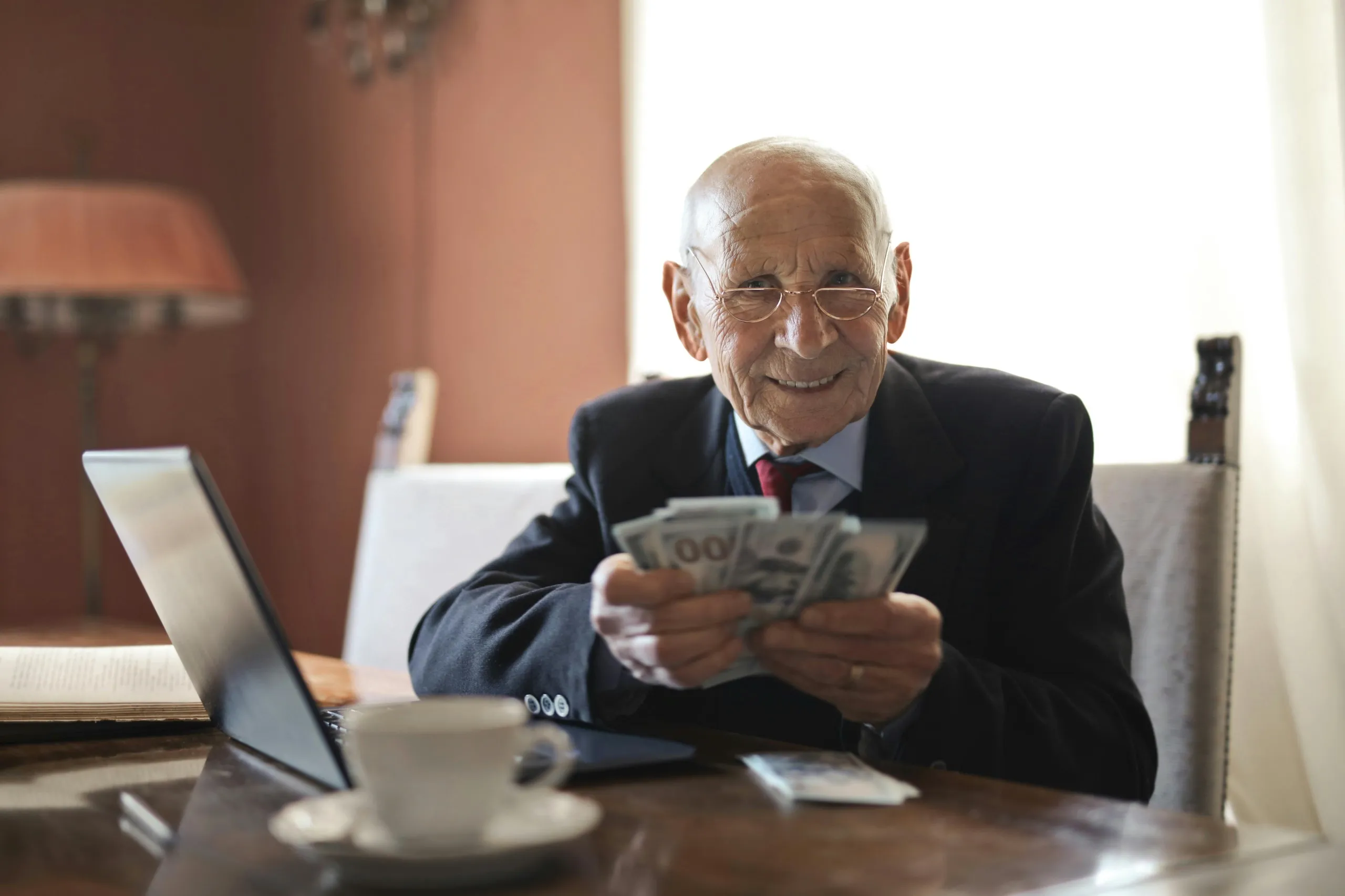 084_01_Should You Pay Your Virtual Assistant Extra When You Delegate Digital Content Creation Tasks Happy elderly man with cash and laptop, showing success from using a virtual assistant for content creation.