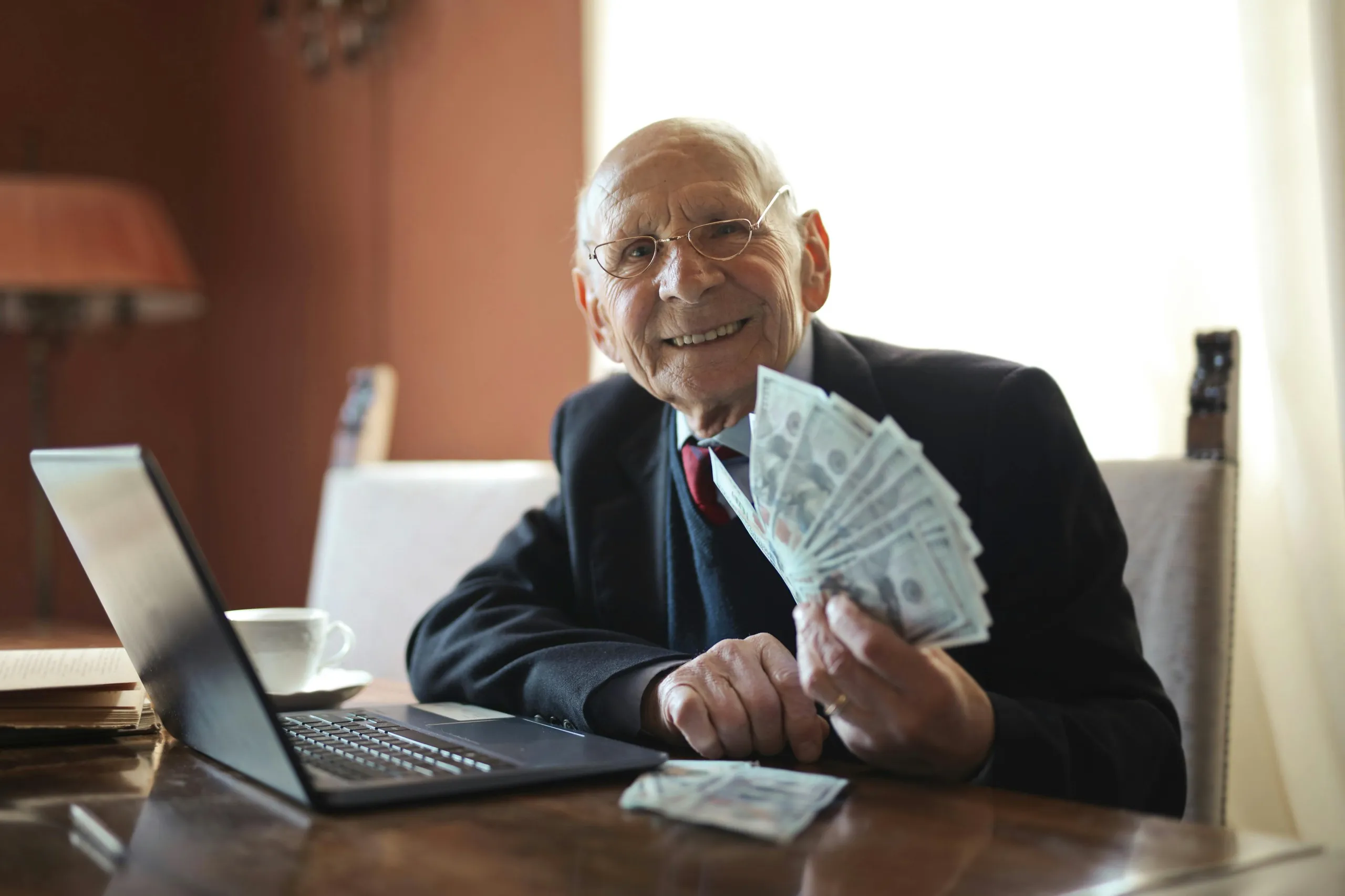 Smiling elderly man holding cash beside a laptop, suggesting earnings from hiring a virtual assistant.