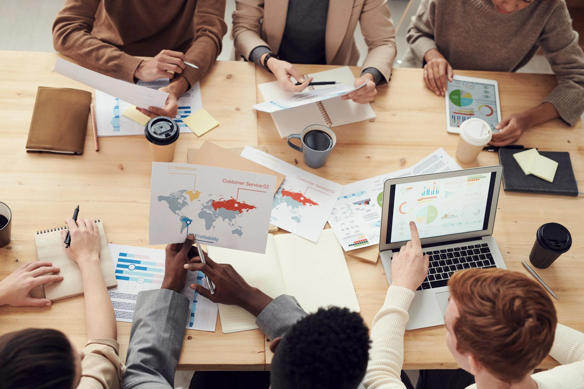 Overhead view of a diverse team collaborating around a wooden table covered in printed analytics charts, laptops, tablets, and coffee cups.