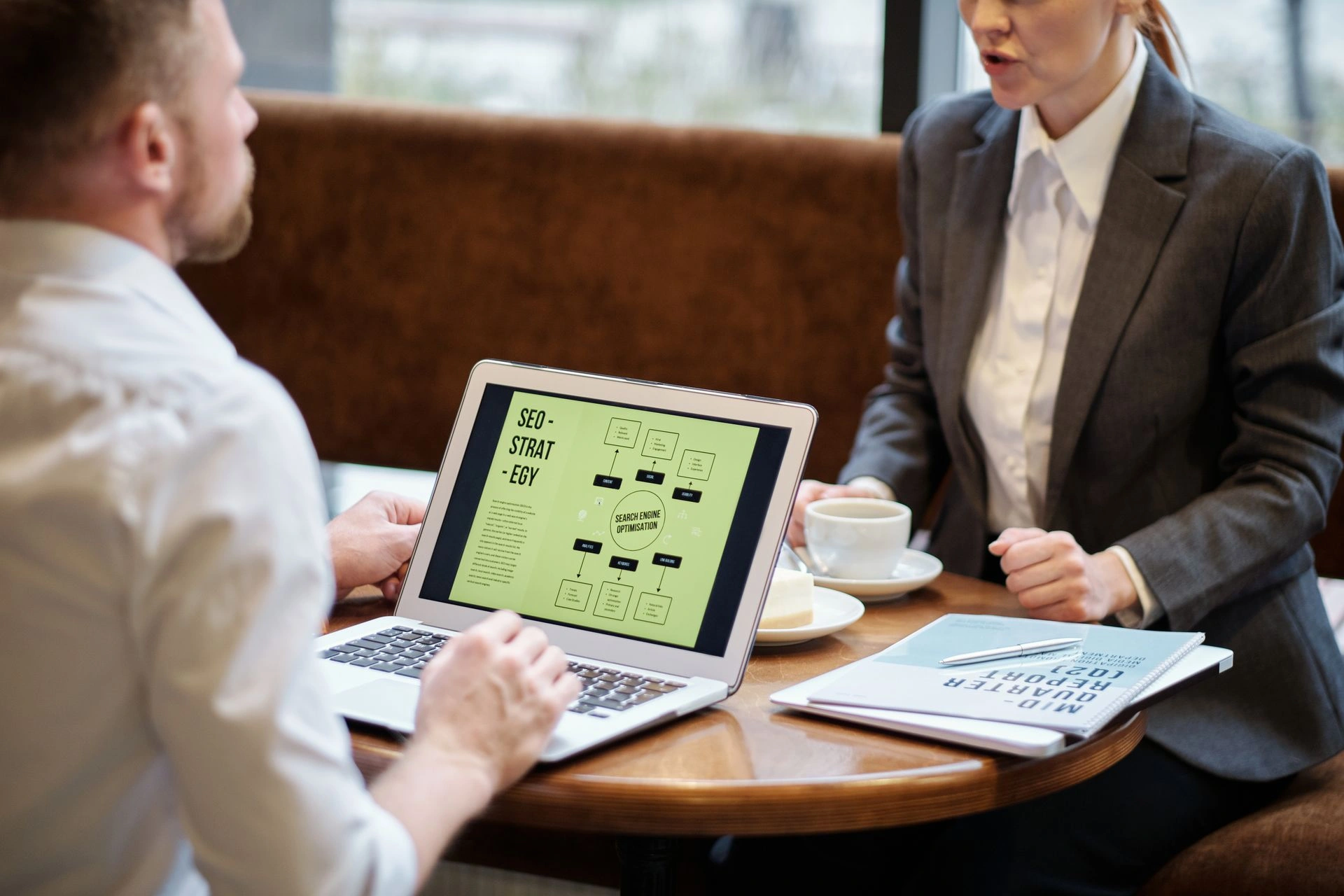 A man and a woman in business suits discussing an "SEO-STRATEGY" flow chart displayed on a laptop screen at a cafe table.