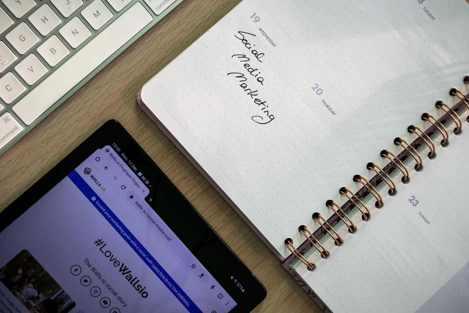 Overhead view of a workspace desk featuring a spiral calendar notebook handwritten with "Social Media Marketing" next to a tablet and computer keyboard.