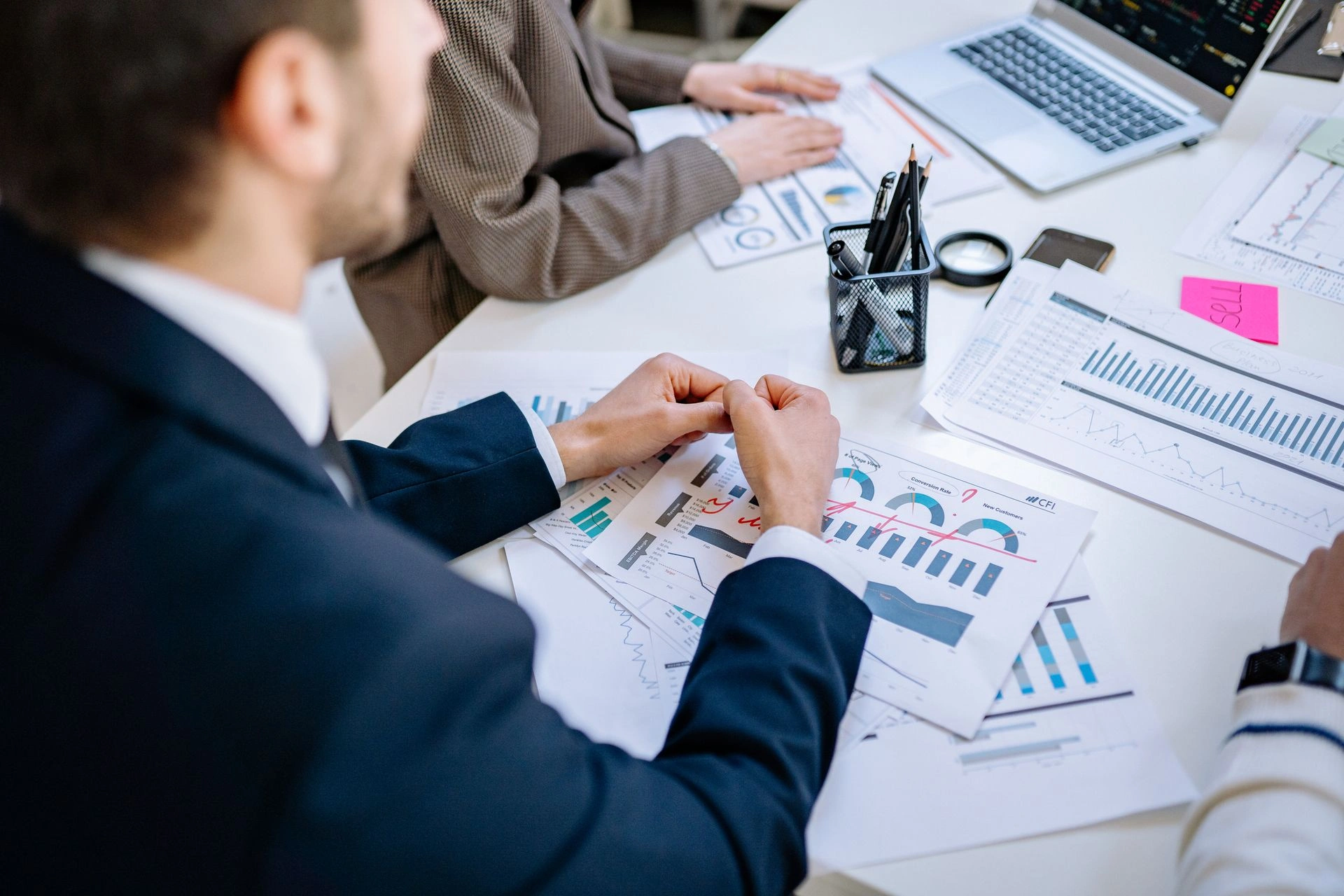 High-angle view of a businessman in a suit leaning over a table covered with financial charts, graphs with red markings, a laptop, and office stationery.