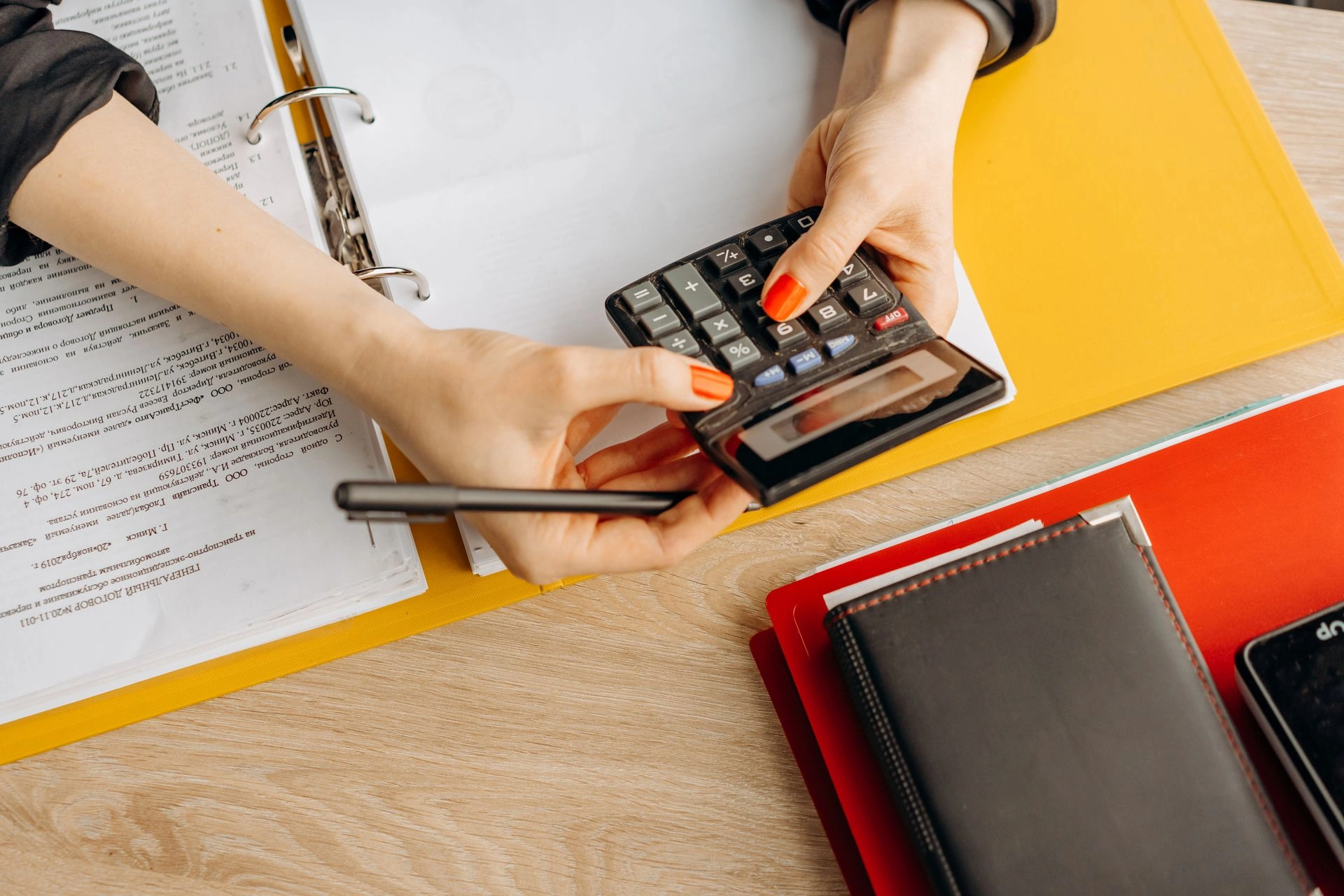 Top-down view of a person with orange-painted nails holding a black calculator while reviewing open document binders and folders on a wooden desk.