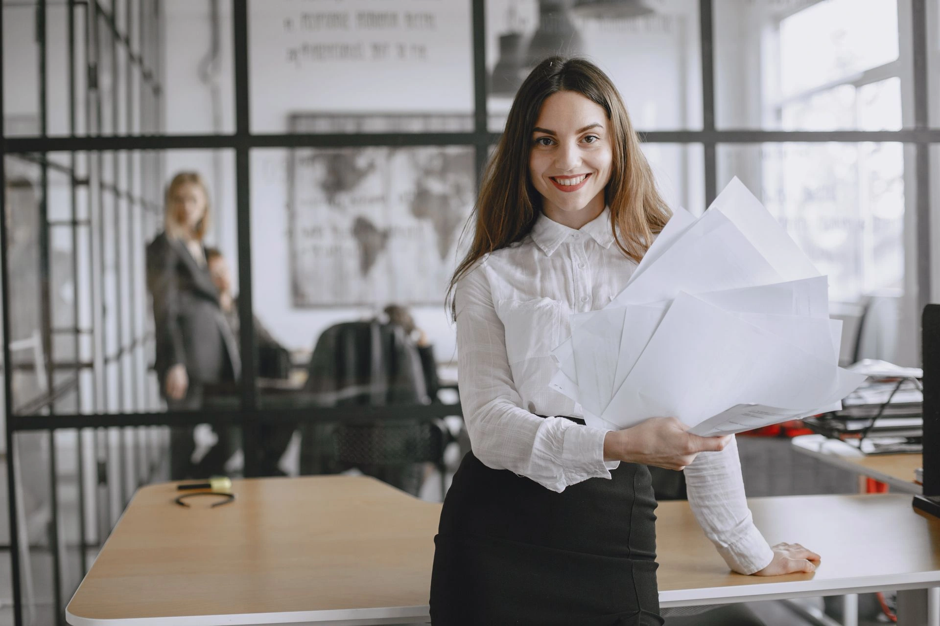 A smiling executive assistant in a white shirt and black skirt holding a stack of papers while leaning on a desk in a modern office.